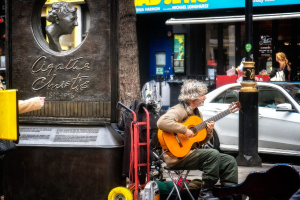 Ein Mann sitzt draußen und spielt eine Akustikgitarre, links daneben eine Statue, im Hintergrund ein Baum und rechts ein weißes Auto und Geschäfte.