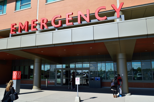 Eine Gruppe von Menschen, die vor einem Notfallgebäude mit Glaswänden und Säulen, einem Namensschild, Hinweistafeln auf Stangen und einem Müllcontainer gehen.