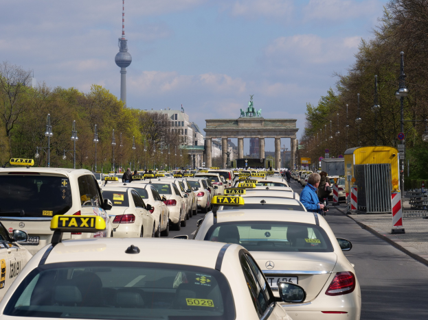 Eine belebte Straße in Berlin, Deutschland, gesäumt von parkenden Taxis, Passanten, Laternenpfählen und Bäumen, mit einem fernen Bogen, Statuen, Turm und einem bewölkten Himmel.