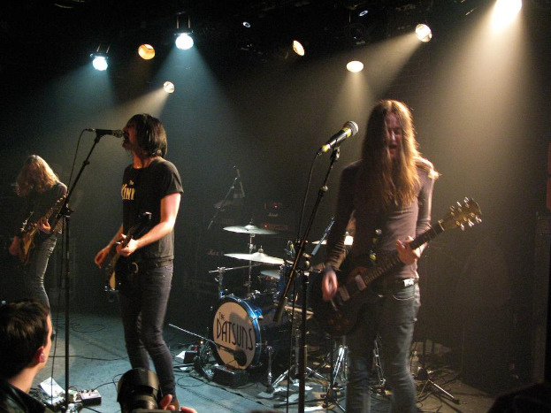 Ein Musikkonzert mit vier Personen: eine filmt, zwei spielen Gitarre und singen, und ein Schlagzeug im Hintergrund.