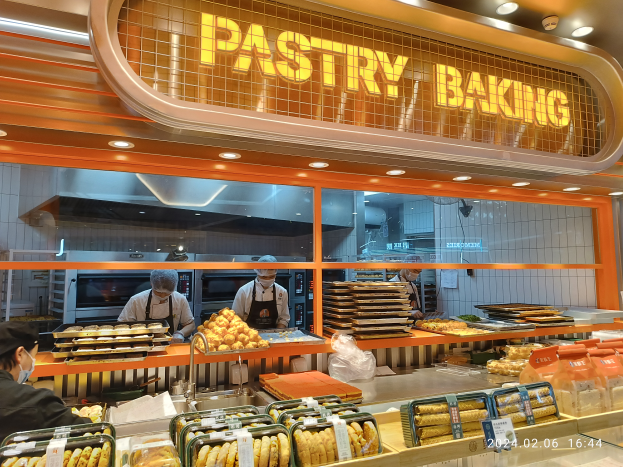 Eine Pasteten-Theke voller verschiedener Lebensmittel, Tabletts und Plastikabdeckungen, mit Personal in Mützen und Masken und einem Namensschild an der Wand unter Deckenbeleuchtung.