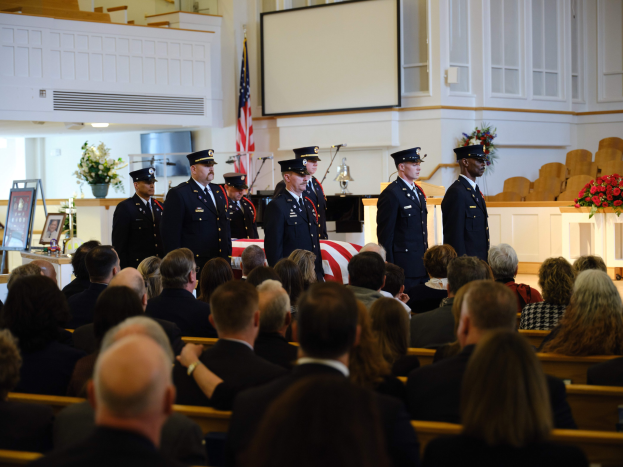 Eine Gruppe uniformierter Personen mit Mützen, die vor sitzenden Trauergästen an einer Beerdigung stehen, mit Blumensträußen, einer Fahne, einem Bildschirm und einer Wand im Hintergrund.