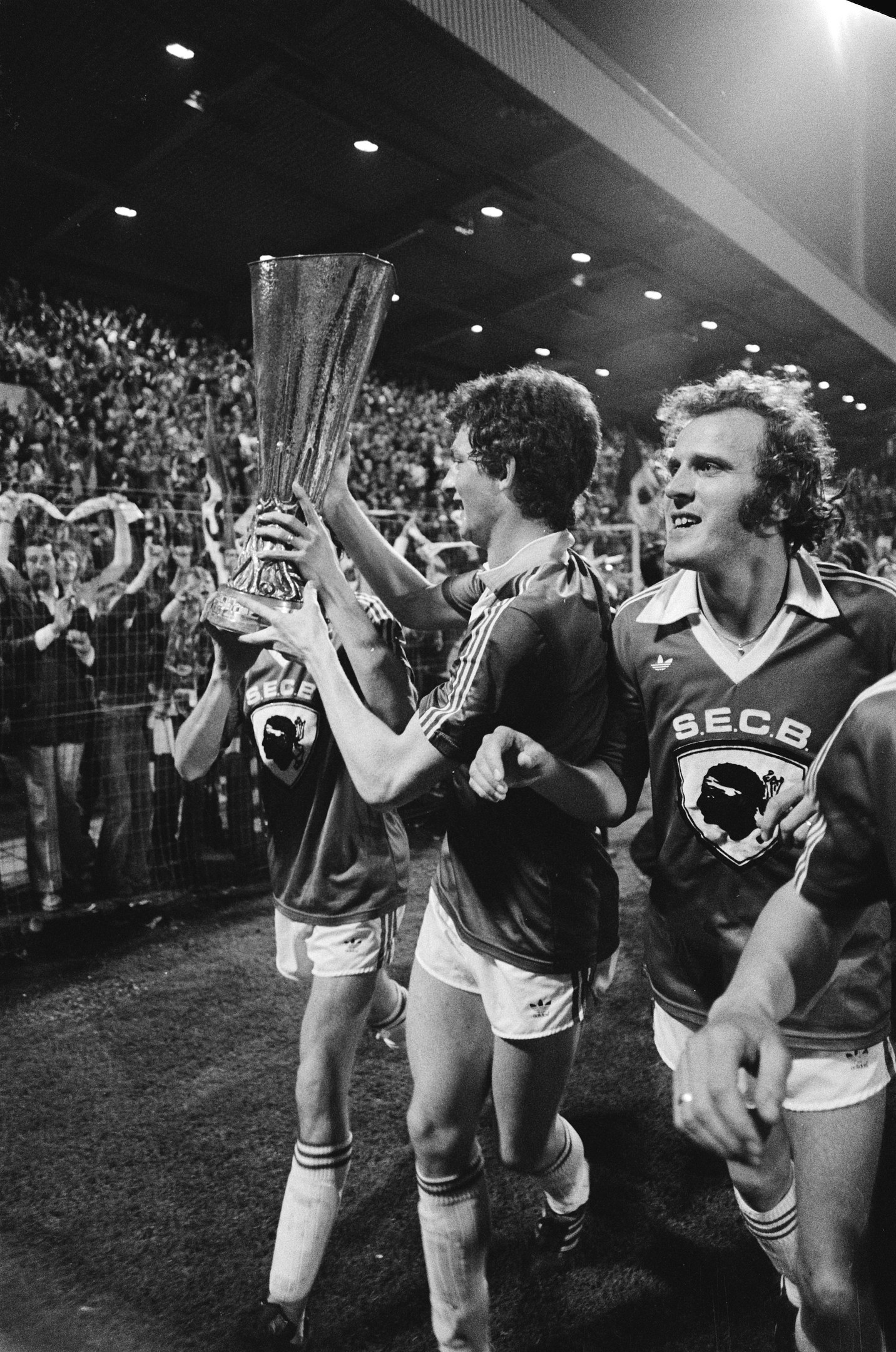 Schwarzes und weißes Bild von Fußballspielern, die auf einem Feld einen Pokal in die Höhe halten, mit Zuschauern im Hintergrund und Scheinwerfern von oben.