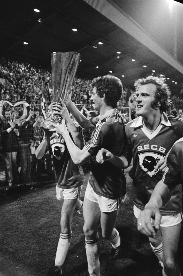 Schwarzes und weißes Bild von Fußballspielern, die auf einem Feld einen Pokal in die Höhe halten, mit Zuschauern im Hintergrund und Scheinwerfern von oben.