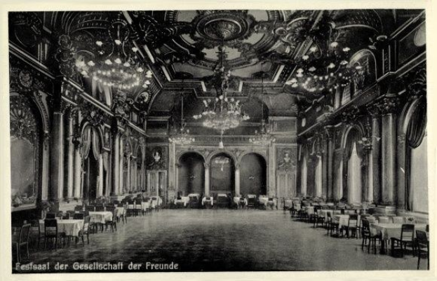Ein altes Schwarz-Weiß-Foto eines großen Raums mit Tischen, Stühlen, Säulen, Bögen und Kronleuchtern an der Decke, mit Text unten, der "Festival der Gesellschaft der Freunde, Berlin" lautet.