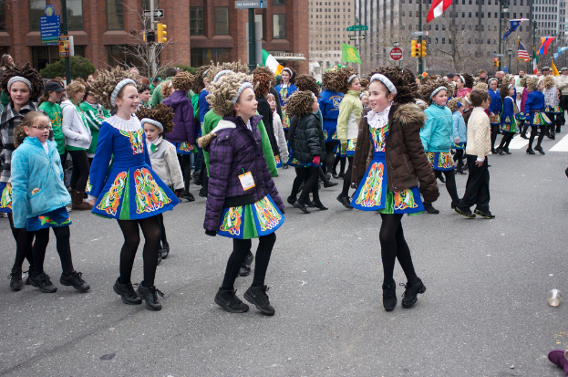 Eine Gruppe von Mädchen in Kleidern mit Jacken und Pullis tanzen auf einer Straße, mit einem roten Gebäude und einem gelben Signalmast dahinter, und Gebäuden und Flaggen an Strommasten im Hintergrund.