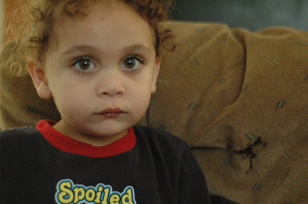 Ein Kind mit lockigen Haaren, das ein schwarzes Shirt mit der gelben Aufschrift 'Spoiled' trägt und vor einem braunen Stoffhintergrund steht.