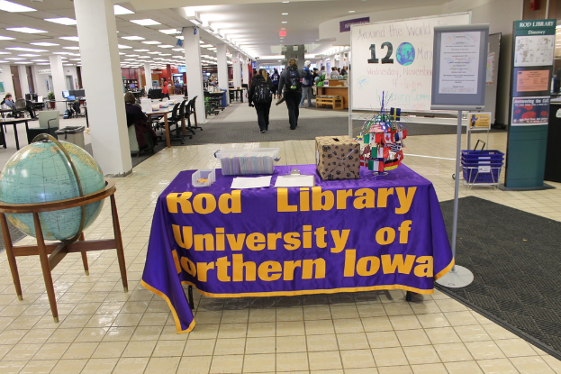 Eine Ausstellung mit einem Tisch, der verschiedene Landesflaggen, eine Box, Papiere und eine Fahne zeigt, eine Kugel links, ein Mitteilungsbrett rechts mit ein paar Menschen in der Nähe und mehrere Tische und Stühle links.