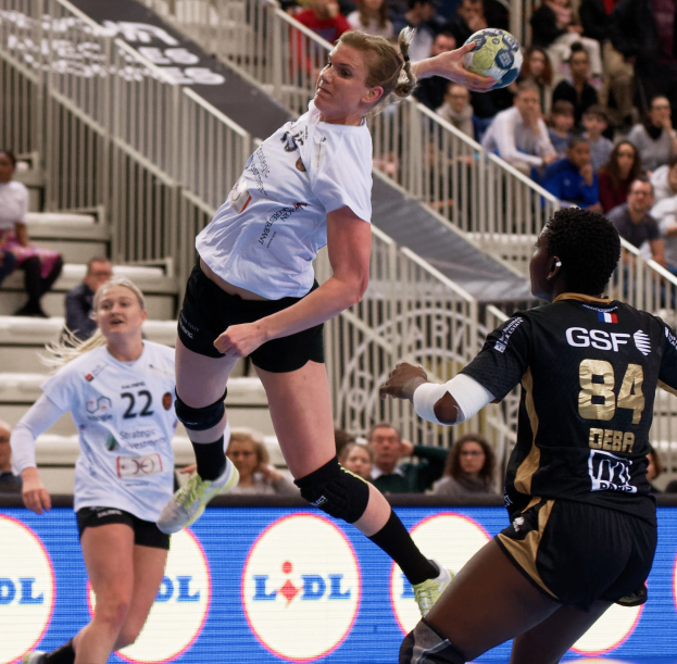 Frauen spielen Handball auf einem Feld mit einem Ball in der Luft, Zuschauern im Hintergrund und einem Schild für die "Handball-Weltmeisterschaft 2019 - Tag 2".