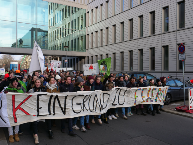 Eine Gruppe von Menschen steht auf einer Straße und hält ein Schild hoch, auf dem "Kein Grind Lo Feuer" steht, im Protest, mit Gebäuden, Bäumen, Pfählen, Schildern, Fahrzeugen und einem klaren blauen Himmel im Hintergrund.