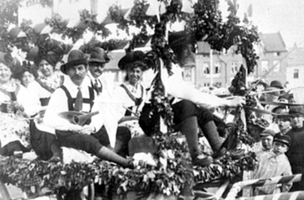 Eine Gruppe von Menschen in traditioneller bayrischer Tracht fährt auf einem mit Blättern geschmückten Festwagen in einem Umzug, einige spielen Musikinstrumente, mit Gebäuden und einem klaren Himmel im Hintergrund, dargestellt in Schwarz-Weiß.