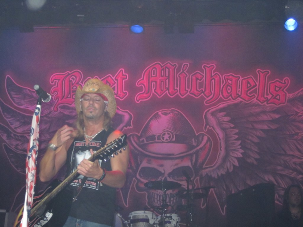 Eine Konzertszene mit einem Gitarristen links neben einem Mikrofon, einer Person in der unteren rechten Ecke, einem Banner im Hintergrund und Drogen in der Mitte unten, mit Licht von oben.