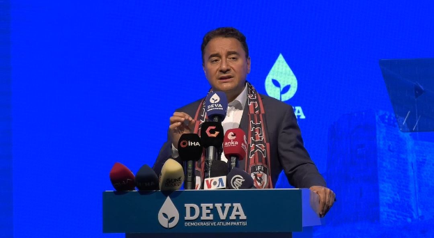 Ein Mann in Mantel und Schal steht hinter einem Pult mit Mikrofonen und spricht hinter einer Tafel mit der Aufschrift "deva-Gesch├Ąftsführer sagt, es sei keine gute Idee für die Zukunft.", mit einem Hintergrundbild und einem Objekt im Hintergrund.