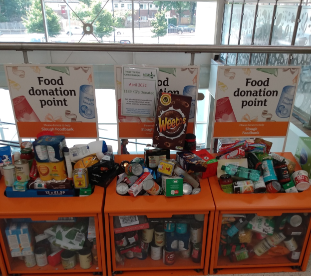 Orangene Schübe mit Lebensmittelspenden gefülllt, darunter Flaschen, Dosen und Kisten, mit informativen Schildern; durch Glasfenster sind Fahrzeuge, Bäume, Gebäude und Himmel zu sehen.