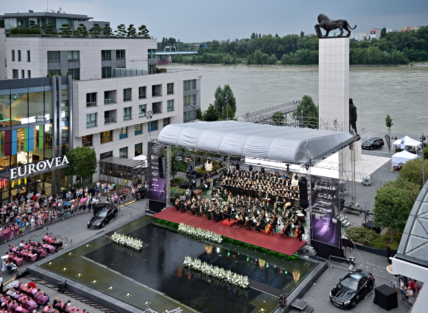 Eine große Menschenmenge auf einer Bühne neben einem Gewässer sitzend, einige halten Musikinstrumente, Fahrzeuge auf der Straße darunter, Banner mit Text, ein Dach mit einem Dach, eine Statue auf einem Pfeiler, Gebäude, Bäume und Laternen im Hintergrund unter einem bewölkten Himmel.