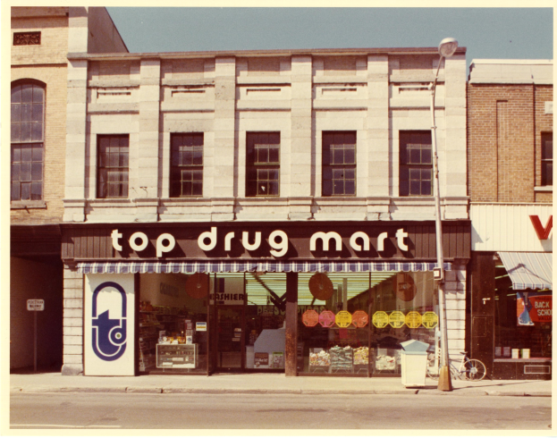 Top-Drogenmart-Filiale an einer Straßenecke mit einem Fahrrad in der Nähe, einem Müllcontainer, einem Straßenpfosten und einem bewölkten Himmel.