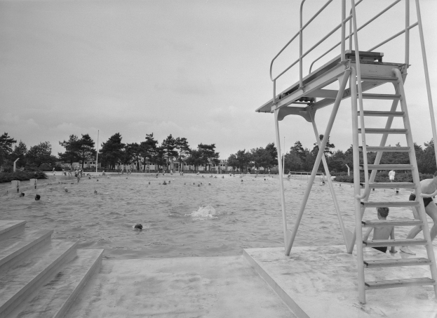 Ein Schwarz-Weiß-Foto von Menschen, die im Wasser an einem Strand schwimmen, mit einem Rettungsturm auf der rechten Seite mit Treppen und Bäumen, Pfählen und einem klaren Himmel im Hintergrund.