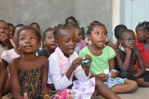 Eine Gruppe trauriger Kinder, die auf dem Boden vor einem Gebäude mit einer Wand und einer Tür im Hintergrund sitzen.