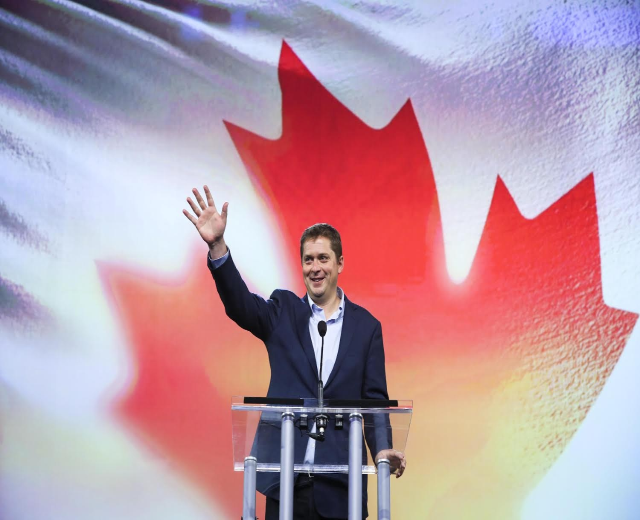 Der kanadische Premierminister Alex Salmond steht an einem Podium mit Mikrofonen, trägt ein Sakko und lächelt, vor einer kanadischen Flagge mit einem Bildschirm im Hintergrund.