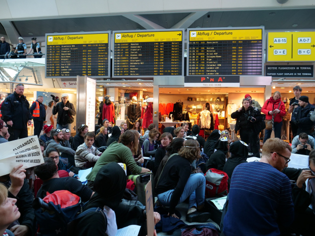 Eine große Gruppe von Menschen versammelt sich in einem Flughafen, einige sitzen mit Taschen und Papieren, andere stehen, mit Texttafeln, Schaufensterpuppen in Kleidern und Deckenlichter im Hintergrund, was auf eine Demonstration hindeutet.