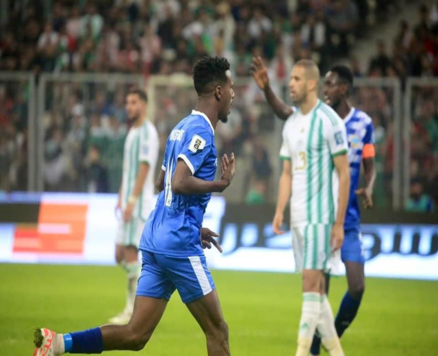 Ein Fußballspieler in einem blauen und weißen Trikot feiert nach einem Tor, umgeben von einer Gruppe von Menschen in weißen und grünen Trikots, mit einem weißen und blauen Brett und Geländer im unscharfen Hintergrund.