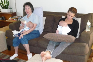 Zwei Frauen auf einer Couch sitzend, die Babys halten, mit Kissen auf der Couch, einer weißen Wand im Hintergrund und einem Schreibtisch mit Büchern und einer Topfpflanze links.