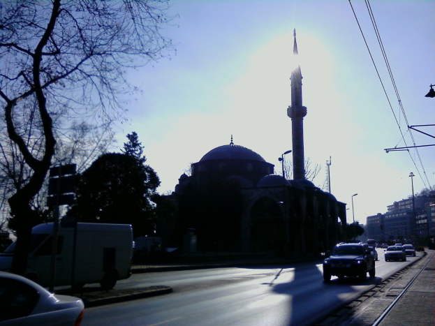 Eine Moschee steht in der Mitte einer Stadtstraße, umgeben von Gebäuden, Straßenlaternen, Strommasten, Stromkabeln, Kraftfahrzeugen, Bäumen und einem klaren blauen Himmel.