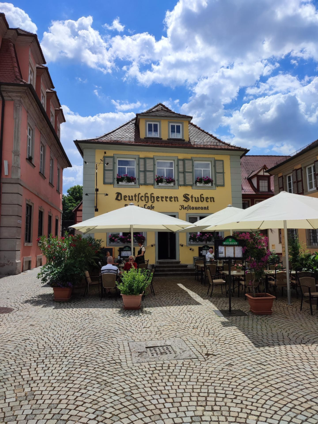 Freisitzbereich des Deutschen Stuben Restaurants in Baden-Württemberg, Deutschland, mit Tischen unter Schirmen, sitzenden und stehenden Menschen und einem gelben Gebäude mit Text darauf unter einem blauen und weißen Himmel.