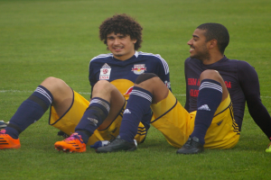 Zwei Fussballspieler sitzen auf dem Boden mit gekreuzten Beinen, tragen Sportbekleidung und schauen sich gegenseitig an.