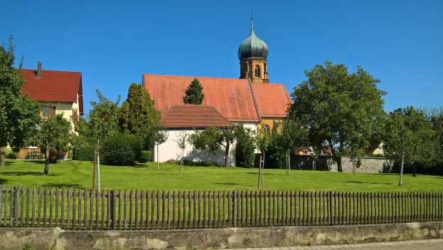 Kirche mit Turm in einer grünen Wiese, umgeben von Häusern, Bäumen, einem Zaun, einem Weg und einer bewölkten Himmel.