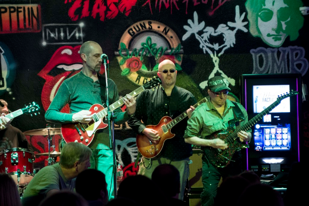 Drei Männer, die auf einer Bühne Gitarren spielen, mit einer Menge darunter und einer bemalten oder gesprayten Wand im Hintergrund.