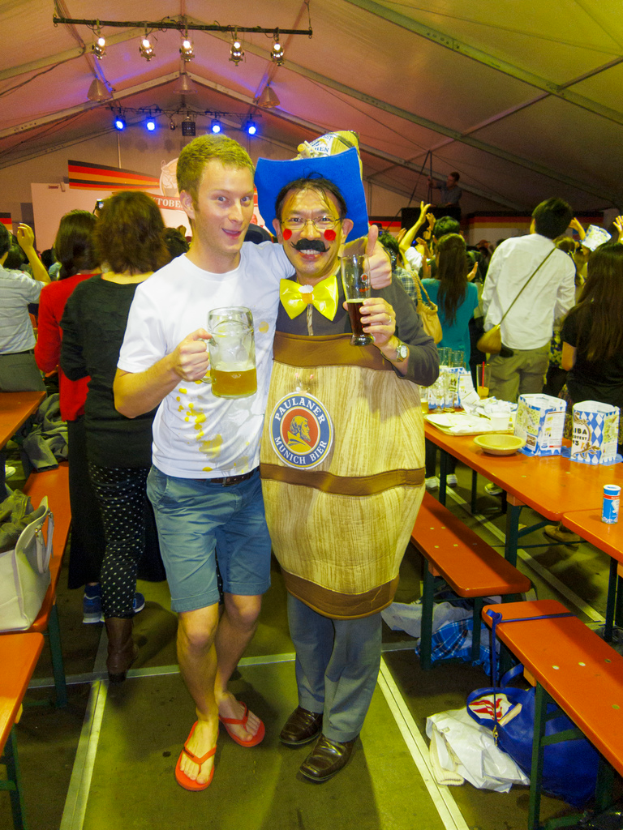 Zwei Personen stehen zusammen, eine hält ein Kind und ein Bier, die andere trägt ein Drama-Kostüm, mit einem Tisch voller Essen in der Nähe und einer Gruppe von Menschen dahinter.