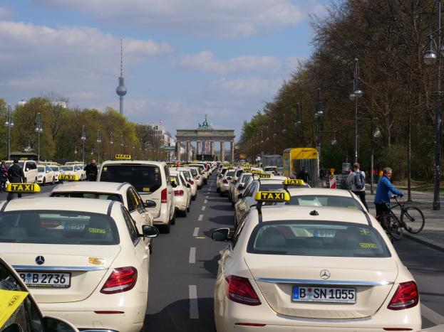 Eine lange Reihe von Taxis, die an einer belebten Straße in Berlin, Deutschland, geparkt sind, mit Fahrradfahrern und Fußgängern auf dem Gehweg, flankiert von Bäumen und Laternen, und Gebäuden, einem Bogen und einem Turm im Hintergrund unter einem bewölkten Himmel.