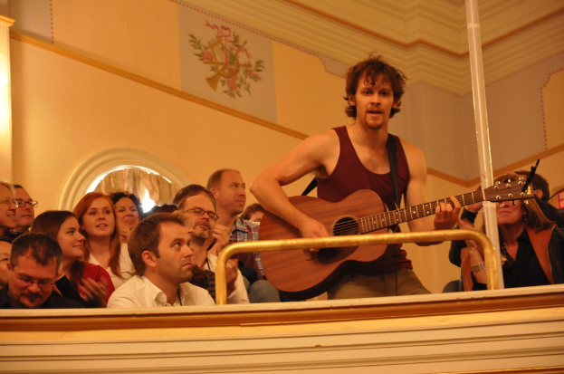 Eine Gruppe von Menschen, die Musik genießen und ein Mann in der Mitte mit einem ärmellosen Hemd singen und eine Gitarre spielen, vor einer braunen Wand mit einem Gemälde oben.