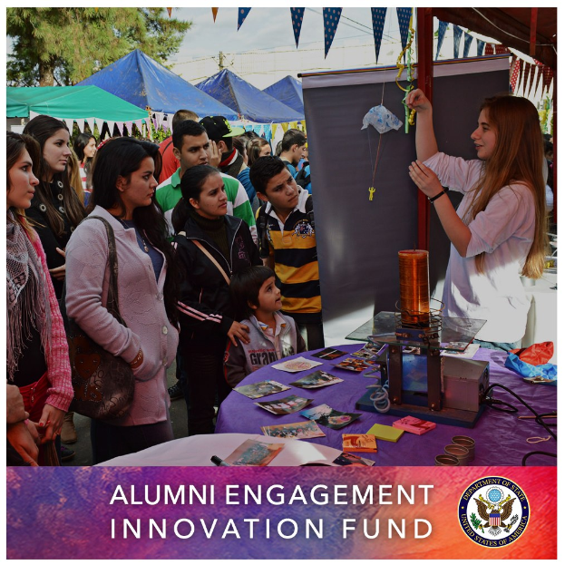 Gruppe von Menschen um einen Tisch versammelt mit einer "Alumni Engagement Innovation Fund"-Aufschrift, umgeben von Gegenständen, mit Zelten, Bäumen und einem klaren blauen Himmel im Hintergrund und Text und ein Logo unten.