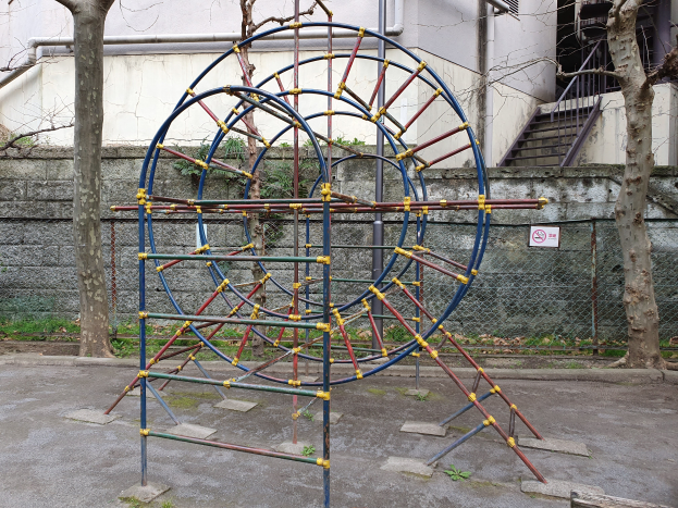 Ein Spielplatz mit einer großen Metallstruktur in der Mitte, umgeben von Bäumen und einer Wand mit Maschendrahtzaun, mit Stufen mit Geländern, die zu einem Gebäude mit Fenstern und Rohren führen, und einem Schild an einem Pfahl, alles auf einem grasbewachsenen Boden.