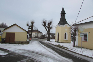 Eine kleine Stadtansicht mit einer zentralen Kirche, umgeben von schneebedeckten Häusern, Bäumen, Schildern, einem Müllcontainer und Stromleitungen unter einem bewölkten Himmel.