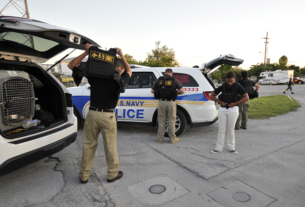 Eine Gruppe uniformierter Polizisten, wahrscheinlich Teil einer Hundestaffel, steht neben einem Polizeiwagen auf einer von Gras, Bäumen, Strommästen mit Drähten und einem Turm umgebenen Straße unter einem klaren Himmel.