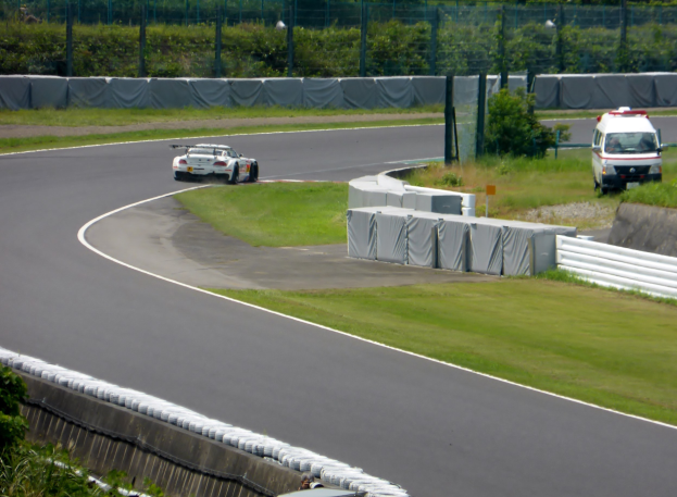 Ein Rennwagen, der auf einer Rennstrecke fährt, mit einem Polizeiauto im Hintergrund, umgeben von Gras, Pflanzen, Bäumen und einem Zaun, mit einem Fahrzeug auf der rechten Seite und Text unten.