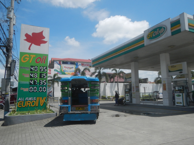 Tankstelle mit einem vor ihr geparkten Lkw, Fahrzeugen auf der Straße, einem Radfahrer, Zapfsäulen, Bäumen, Gebäuden, Wolken und einem Strommast mit Drähten im Hintergrund.