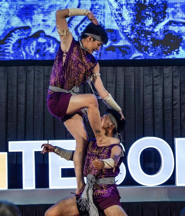 Zwei Akrobaten führen auf der Bühne eine Darbietung auf, wobei der Mann die Frau in seinen Armen hält, während beide purpurne Kleider tragen, mit einem schwarzen Vorhang und einer Leinwand im Hintergrund und sitzenden Zuschauern darunter.