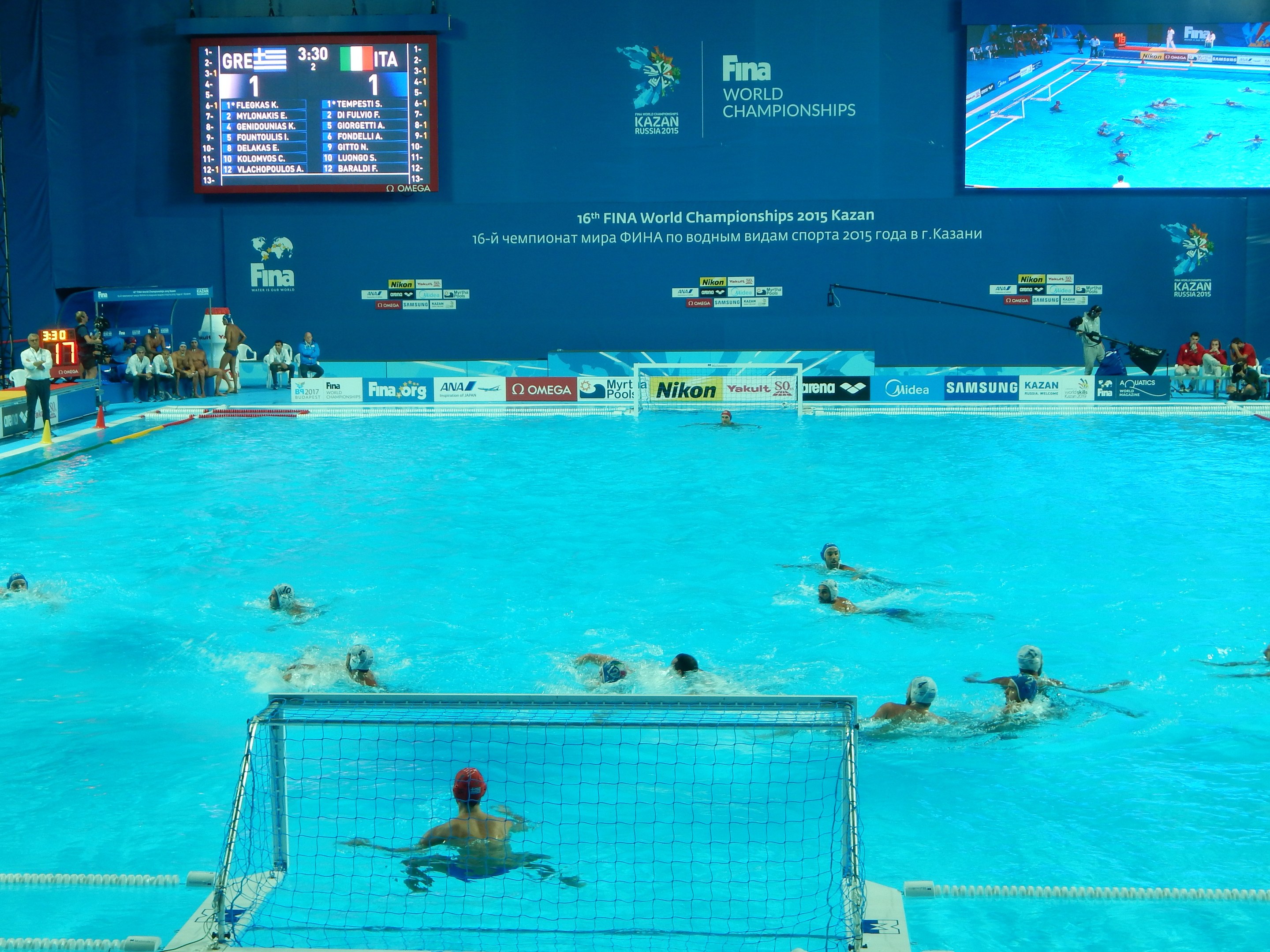 Wasserballspieler bei den 2016 FINA-Weltmeisterschaften in Kazan im Pool gegeneinander antretend, mit Zuschauern auf der linken Seite und einem werbebeschrifteten Bildschirm im Hintergrund.