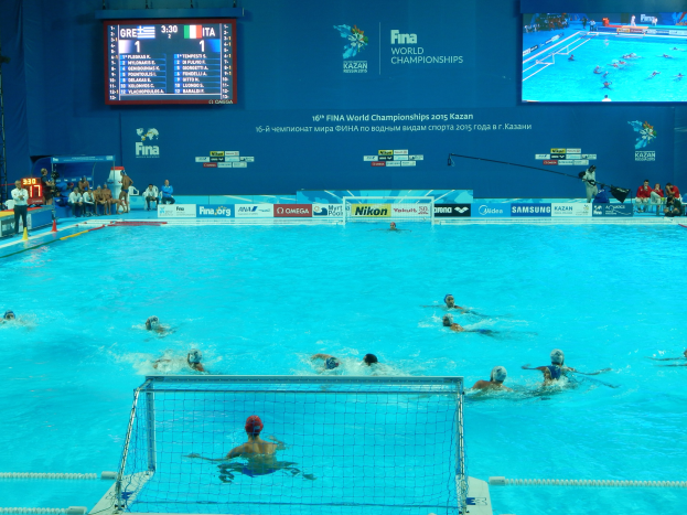 Wasserballspieler bei den 2016 FINA-Weltmeisterschaften in Kazan im Pool gegeneinander antretend, mit Zuschauern auf der linken Seite und einem werbebeschrifteten Bildschirm im Hintergrund.