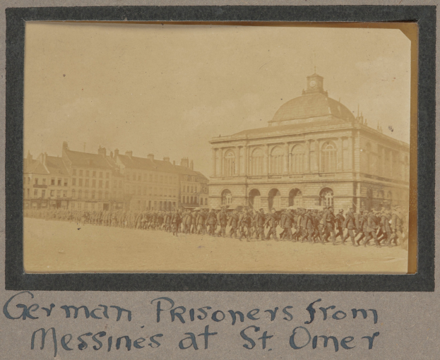 Eine Gruppe deutscher Gefangener aus Messines steht vor einem Gebäude mit einem klaren blauen Himmel im Hintergrund, und Text ist unten im Bild sichtbar.