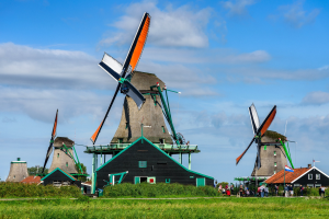 Gruppe von Windmühlen in Kinderdijk, Niederlande, mit Menschen in der Nähe, umgeben von grünem Gras und Pflanzen, Bäumen im Hintergrund und einem klaren blauen Himmel.