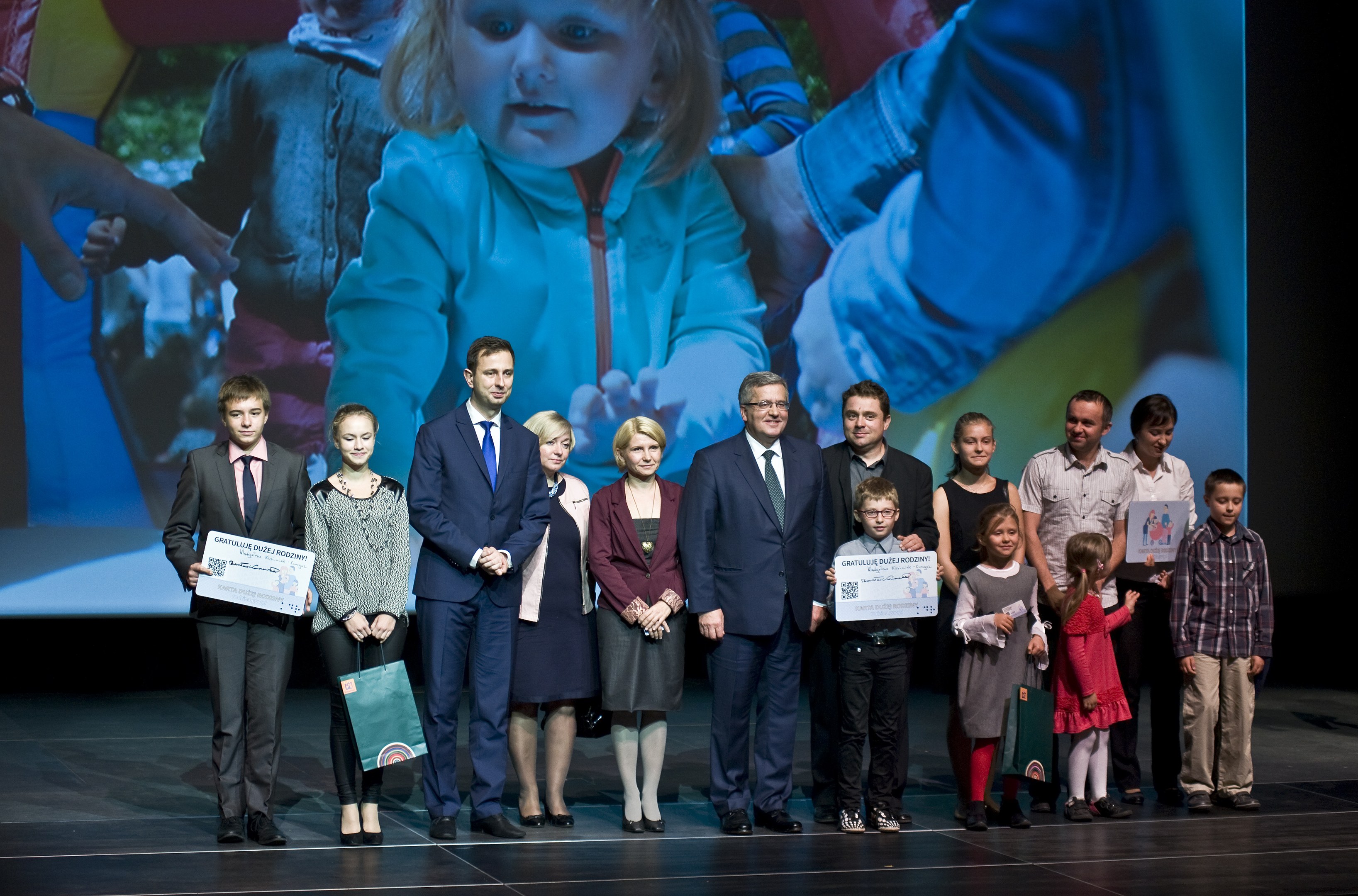 Gruppe von Menschen auf einer Bühne mit Taschen und Urkunden, hinter ihnen ein Bildschirm mit einem Bild eines Kindes und anderen im Hintergrund.
