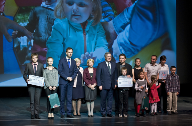 Gruppe von Menschen auf einer Bühne mit Taschen und Urkunden, hinter ihnen ein Bildschirm mit einem Bild eines Kindes und anderen im Hintergrund.