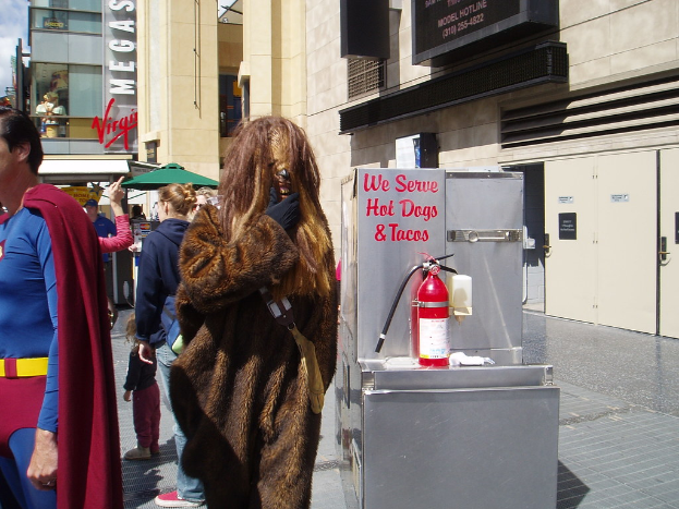 Eine Menschenmenge, darunter einige in Kostümen, versammelte sich vor Gebäuden mit sichtbarer Beschriftung, einer Box mit Schrift und einem Feuerlöscher.