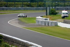 Ein Rennwagen fährt auf einer Rennstrecke mit einem Polizeiwagen im Hintergrund, umgeben von Gras, Pflanzen, Bäumen und Absperrungen, mit einem Fahrzeug auf der rechten Seite und Text unten.