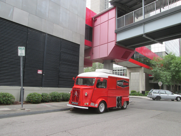 Ein Van steht in der Mitte des Bildes mit einem Auto auf der rechten Seite, und Gebäude sind im Hintergrund zu sehen.